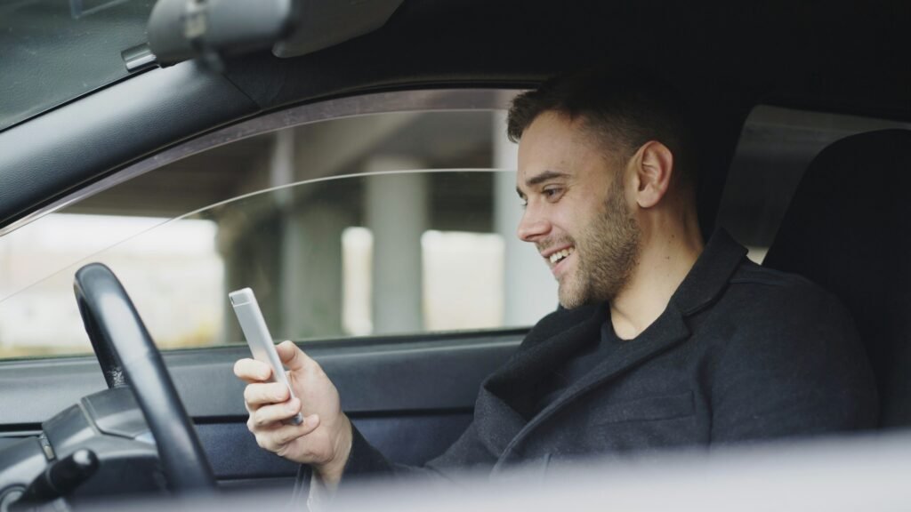 Smiling driver holding a phone in his car, representing safe and insured side hustles using a car.